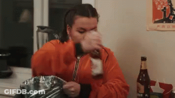 Woman In Orange Jacket Drinking From Bottle While Cooking GIF