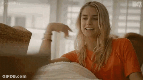 Woman In Orange Shirt Sitting On Couch Hand On Chin GIF