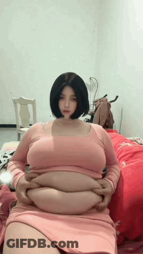 Woman In Pink Dress Sitting On Bed Hands On Stomach GIF