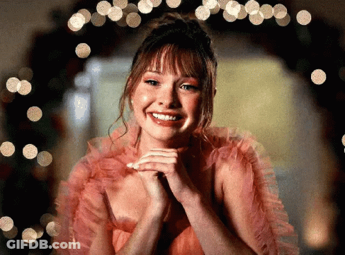 Woman In Pink Dress Smiling In Front Of Christmas Tree GIF