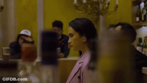 Woman In Pink Jacket Sitting At Table In Front Of Yellow Wall GIF