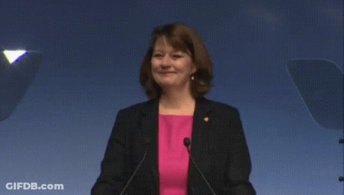 Woman In Pink Shirt And Black Jacket At Podium With Microphones GIF