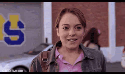 Woman In Pink Shirt And Denim Jacket Smiling In Front Of Building With Letter S GIF