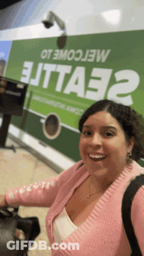 Woman In Pink Sweater Smiling In Front Of Green Sign Saying Elitta32 GIF
