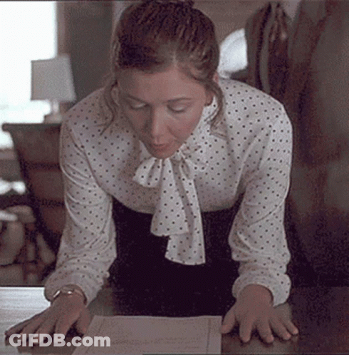 Woman In Polka Dot Blouse Kneeling At Desk Looking At Paper GIF