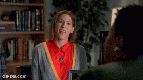 Woman In Red Shirt And Grey Sweater Waving At Another Woman In Front Of Bookshelf GIF
