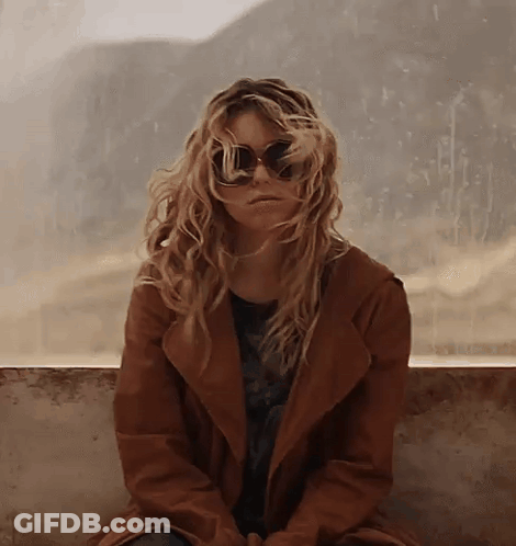 Woman In Sunglasses And Brown Coat Sitting On Wall GIF