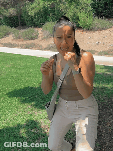 Woman In Tan Tank Top And White Pants Kneeling In Grass GIF