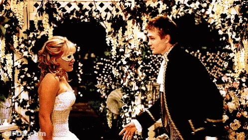 Woman In White Dress And Mask Next To Man In Black Suit GIF