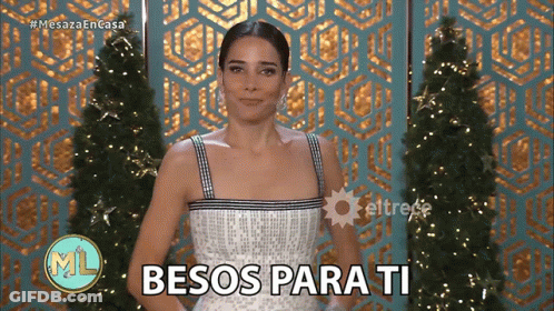 Woman In White Dress Says Besos Para Ti In Front Of Christmas Tree GIF