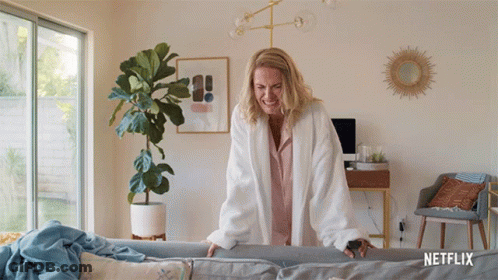 Woman In White Robe Standing Next To Couch With Netflix Logo GIF