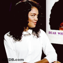 Woman In White Shirt Sitting In Front Of Dear White Sign GIF