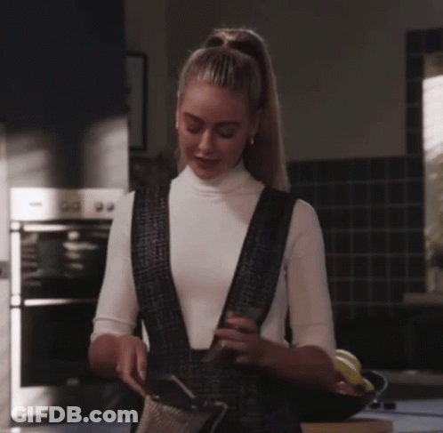 Woman In White Turtleneck And Black Overalls In Kitchen GIF
