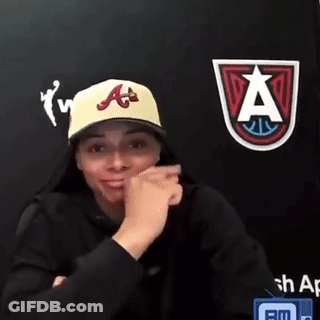 Woman In Yellow Hat And Black Hoodie Sitting In Front Of Wall With Logo GIF