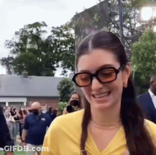 Woman In Yellow Shirt And Sunglasses Smiling At Crowd GIF
