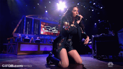 Woman Kneeling Singing Into Microphone In Front Of Dolly Parton Screen GIF