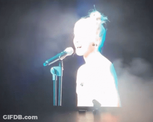 Woman Singing Into Mic On Stage With Screen Behind GIF