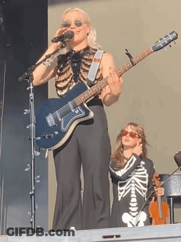 Woman Singing Into Microphone Holding Guitar Beside Girl In Skeleton Outfit GIF