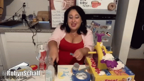 Woman Sitting At Table In Front Of Fridge With Robyn Schall Sign GIF
