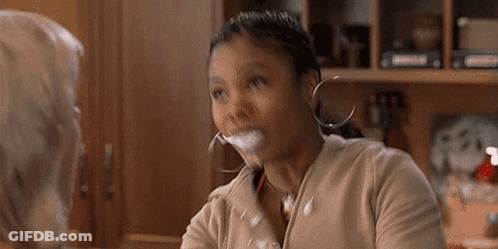 Woman Sitting At Table With Glass Of Milk In Mouth Talking To Another Woman GIF