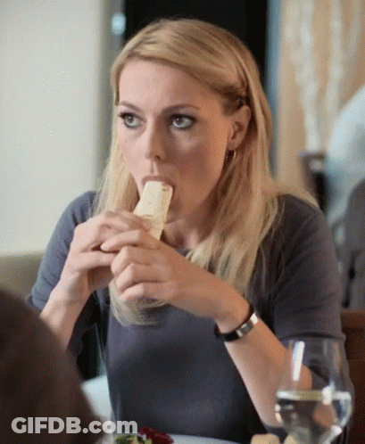Woman Sitting At Table With Sandwich In Mouth GIF