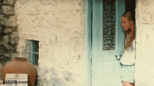 Woman Standing In Front Of Blue Door With Boxes Saying Attar GIF