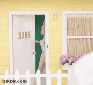 Woman Standing In Front Of Yellow House Behind White Picket Fence GIF