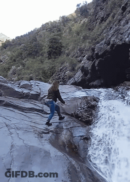 Woman Walking Down Rocky Cliff Near Waterfall GIF