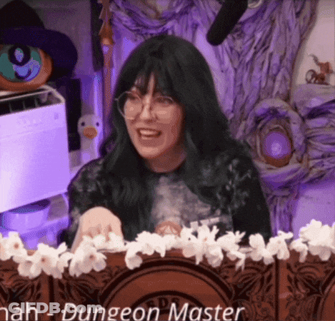 Woman With Green Hair And Glasses At Table With Flowers And Microphone GIF