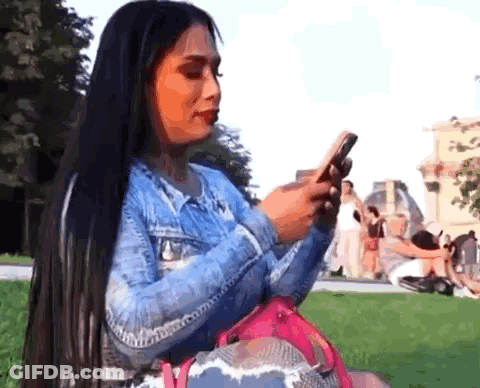 Woman With Long Black Hair Sitting In Grass Looking At Phone GIF