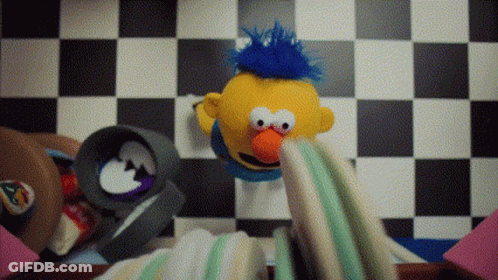 Yellow Puppet With Blue Mohawk Sitting On Checkered Floor GIF