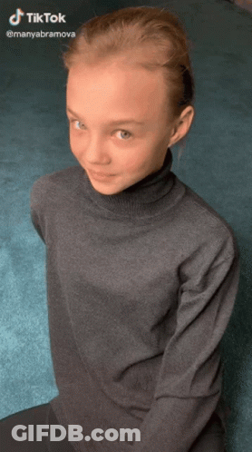 Young Girl In Turtleneck Sitting On Floor Looking At Camera GIF