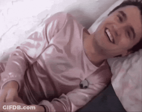 Young Man In Pink Shirt Laying On Bed Smiling GIF