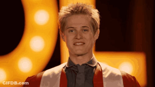 Young Man In Red And White Robe Smiling In Front Of Yellow Sign GIF