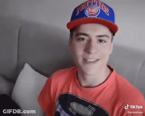 Young Man In Red Shirt And Blue Hat Sitting On Couch GIF