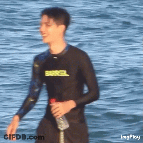 Young Man In Wetsuit Walking On Beach Holding Water GIF