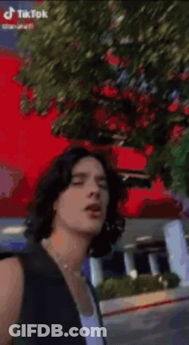 Young Man With Long Hair And Pearl Necklace In Front Of Red Flower GIF