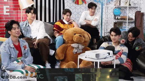 Young Men Sitting Around Teddy Bear And Projector GIF