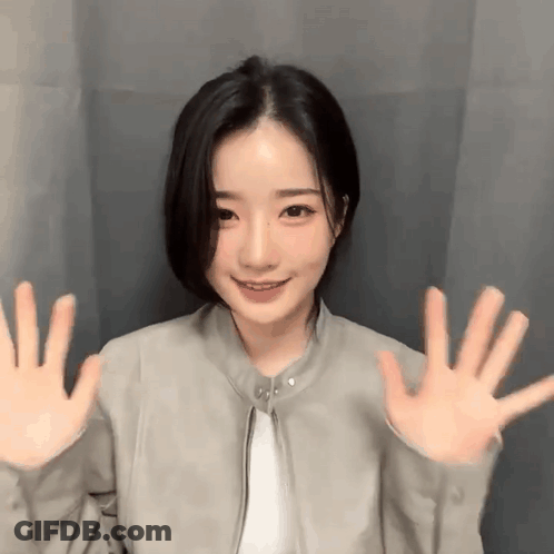 Young Woman In Gray Jacket Waving Hands By Curtain GIF