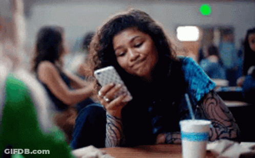 Young Woman Sitting At Table Looking At Cell Phone GIF