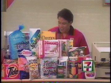 A 7 Eleven Cashier Stands Behind A Counter Full Of Groceries Including Cheerios GIF