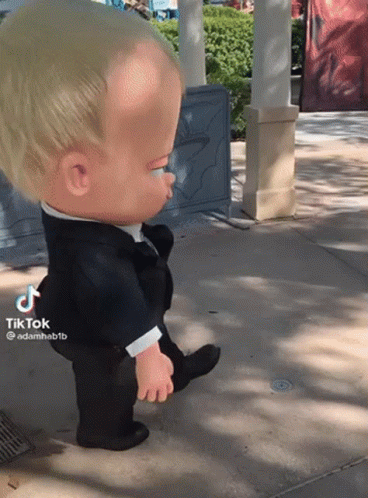 A Baby Doll In A Tuxedo Is Walking On A Sidewalk GIF