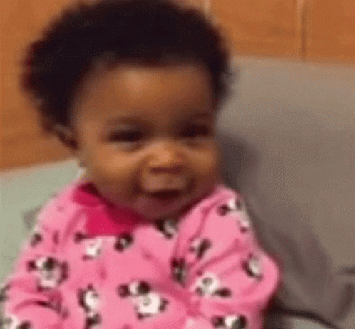 A Baby Girl In A Pink Pajama Set Is Smiling While Sitting On A Bed . GIF