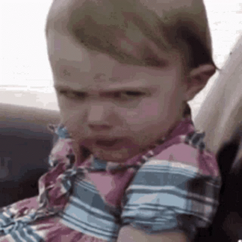 A Baby Girl Is Sitting In A Car Seat With A Serious Look On Her Face . GIF