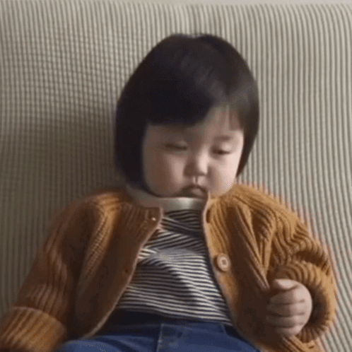 A Baby Girl Is Sitting On A Couch Wearing A Sweater And Striped Shirt . GIF