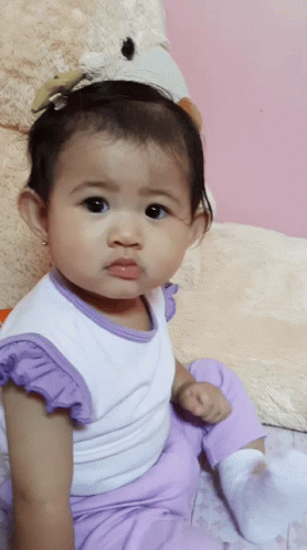 A Baby Girl Wearing Purple Pants And A White Shirt Is Sitting Next To A Teddy Bear GIF