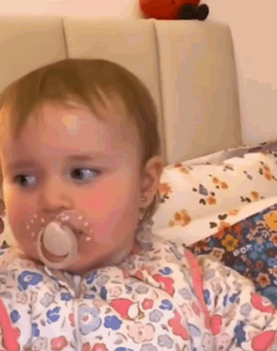 A Baby Girl With A Pacifier In Her Mouth Is Sitting On A Bed . GIF
