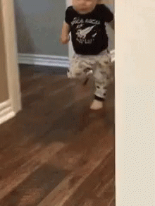 A Baby In A Black Shirt Is Walking On A Wooden Floor GIF