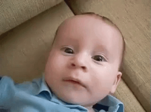 A Baby In A Blue Shirt Is Laying On A Couch Making A Funny Face . GIF