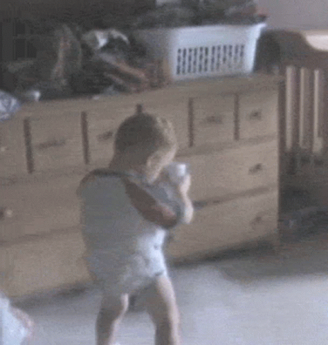 A Baby In A Diaper Is Standing In Front Of A Dresser With A Basket In The Background GIF